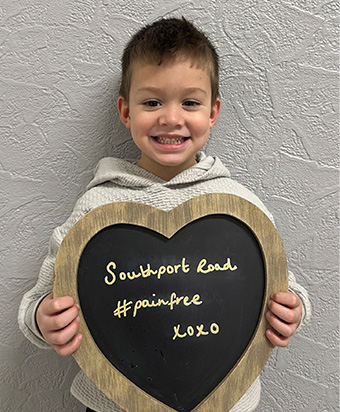 A young patient holding a heart-shaped board reading "Southport Road #painfree"