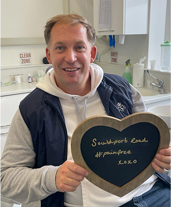 A smiling patient holding a heart-shaped board reading "Southport Road #painfree"